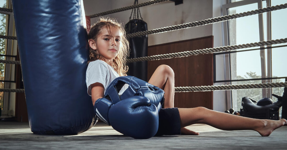 Little tired girl with blue helmet is resting on the ring next to punching bag