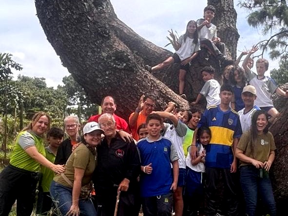Grupo de niños y adolescentes reunidos alrededor de un gran árbol, en un entorno natural y verde