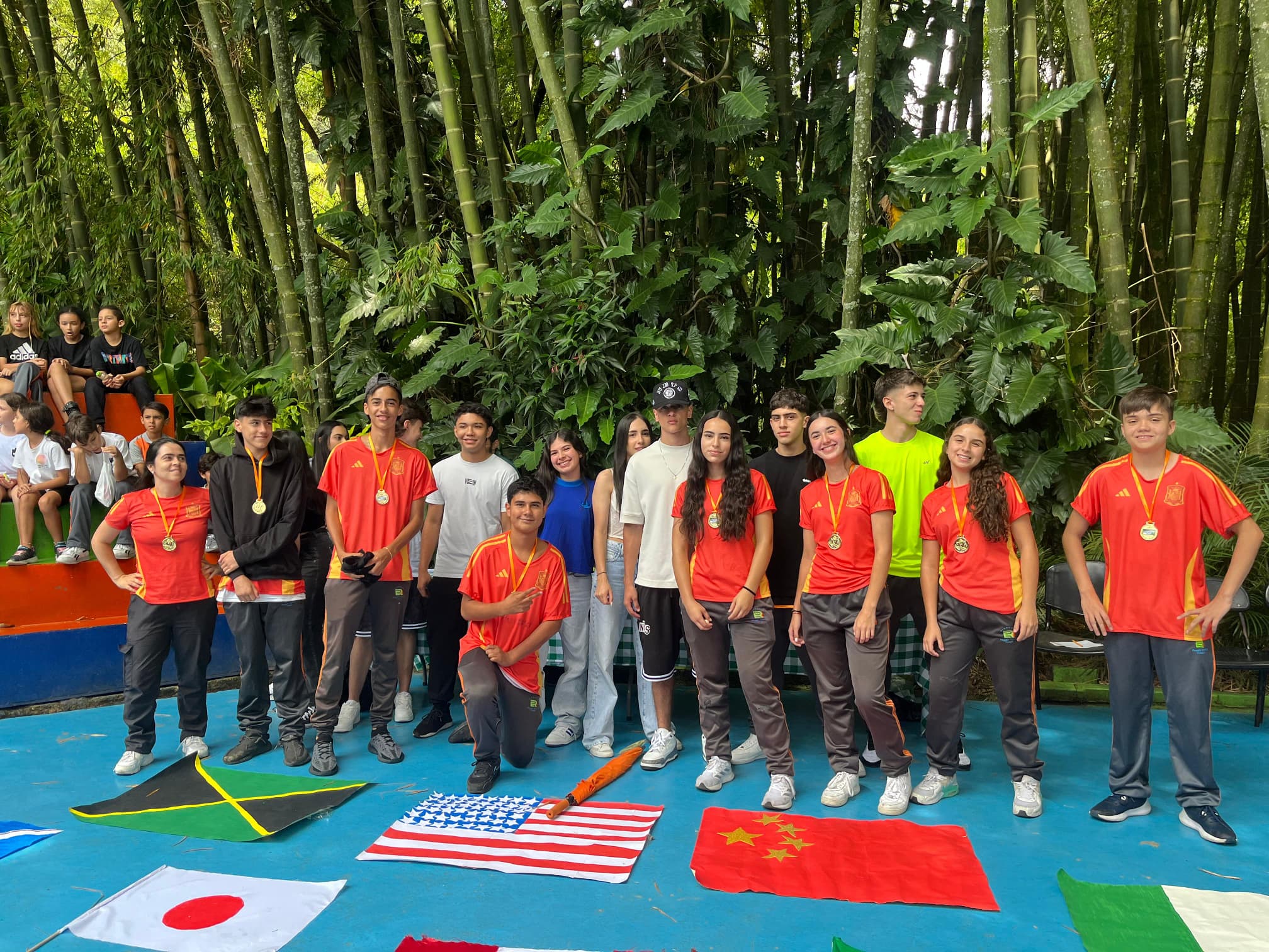 A group of young students, some wearing medals, stand in front of bamboo, with various country flags displayed on the ground in front of them. Other groups are seated in the background.